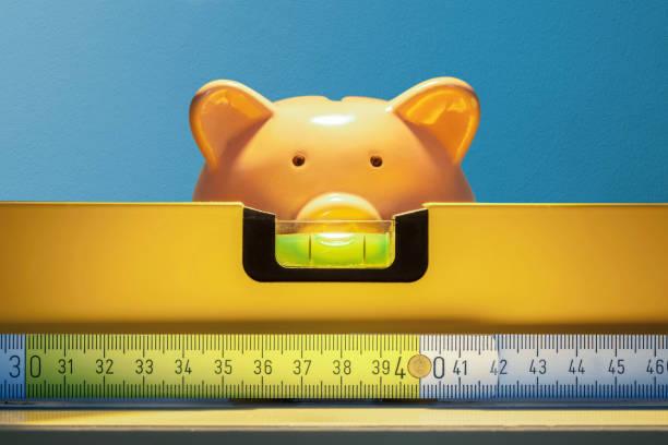 Piggy bank looks over the bubble level on the shelf against blue wall Piggy bank looks over the bubble level on the shelf against blue wall front home extension costs stock pictures, royalty-free photos & images